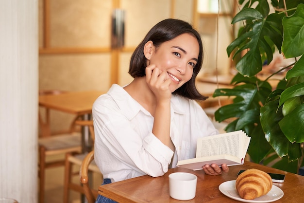 Kostenloses Foto romantische asiatische frau, die mit buch im café sitzt und croissants isst und kaffee trinkt, liest und lächelt