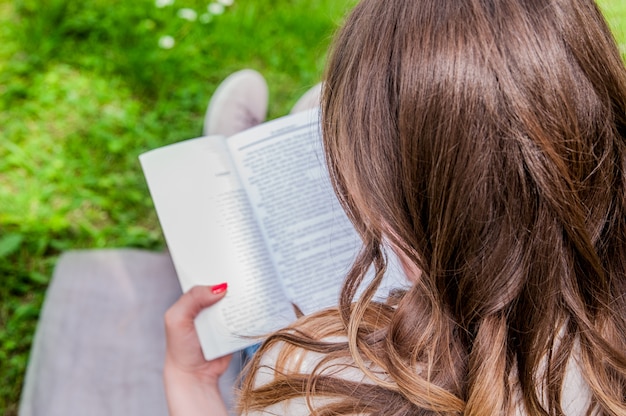 Kostenloses Foto schöne junge brünette sitzt auf einem frischen frühling in einem park, ein buch zu lesen. nahaufnahme einer schönen jungen frau liest buch im park. frau, die ein buch liest, das draußen liest