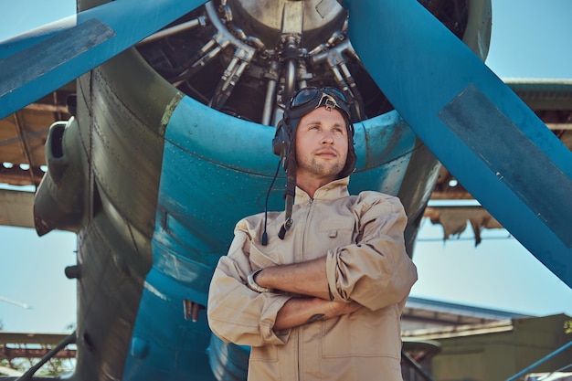 Kostenloses Foto schöner pilot in voller flugausrüstung, der mit verschränkten armen in der nähe von militärflugzeugen steht.