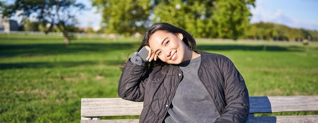 Kostenloses Foto schönheitsporträt eines lächelnden asiatischen mädchens mit dunklen kurzen haaren, das auf einer bank im park sitzt und zuschaut