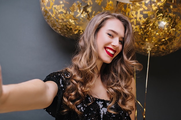 Kostenloses Foto selfie-porträt der jungen charmanten frau mit den roten lippen, dem langen brünetten haar, das mit großen luftballons voll mit goldenen lametta lächelt. positivität ausdrücken, party feiern.