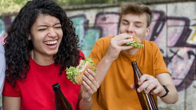 Smiley-Teenager, die Burger im Freien mit Getränk essen