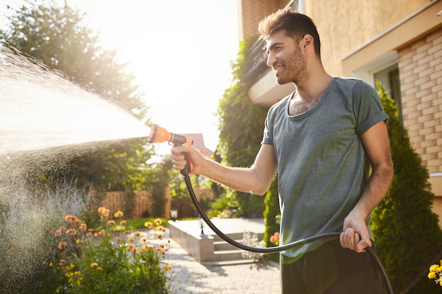 Kostenloses Foto sommermorgen im landhaus. porträt des jungen attraktiven hellbraunen bärtigen mannes im blauen t-shirt lächelnd, wasserpflanzen mit schlauch gießend, im garten arbeitend.