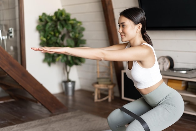 Kostenloses Foto süßes asiatisches fitness-mädchen zu hause, das workout-kniebeugen mit dehnbarem elastischem seil an ihren beinen macht ...