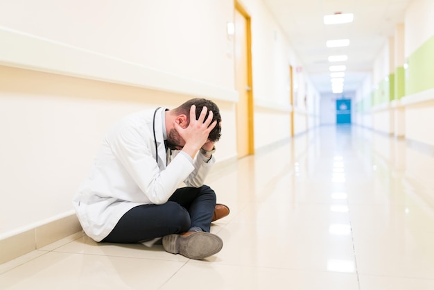 Kostenloses Foto trauriger arzt in der mitte des erwachsenen mit kopf in den händen, der auf dem boden gegen die wand im korridor im krankenhaus sitzt