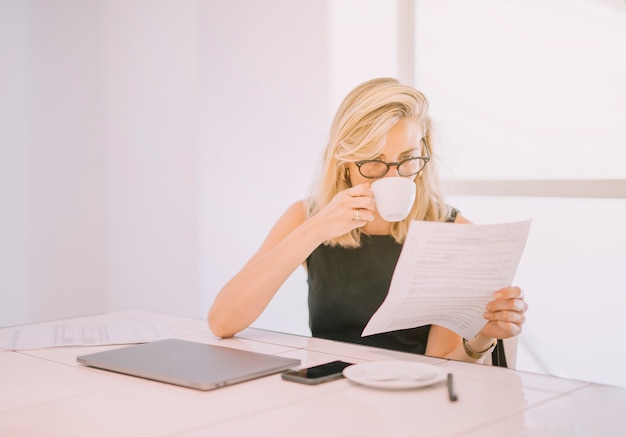 Kostenloses Foto trinkender kaffee der jungen geschäftsfrau beim lesen des dokuments im büro