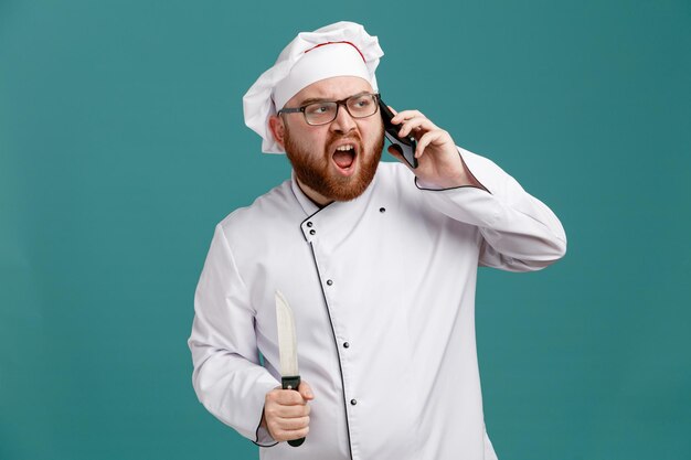 Unzufriedener junger männlicher Koch mit einheitlicher Brille und Mütze mit Messer, der auf die Seite blickt, die am Telefon isoliert auf blauem Hintergrund spricht