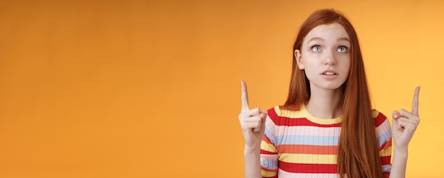 Kostenloses Foto verblüfft begeistert junge rothaarige frau peer konzentriert zeigt nach oben zeigefinger nach oben schauen konzentriert aufgeregt halten atem amüsiert leistung stehend orange hintergrund fasziniert und neugierig