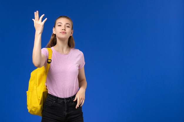 Vorderansicht der Studentin im rosa T-Shirt mit gelbem Rucksack an der hellblauen Wand