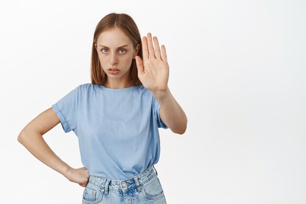 Wütende blonde Frau streckt die Hand aus und runzelt die Stirn, sagt nein, stimme nicht zu, lehne ab oder lehne ab, verbiete Maßnahmen, stimme nicht mit der Person überein, steht über weißer Wand