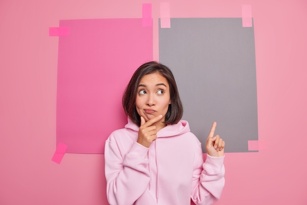 Kostenlose Foto zögernde brünette junge asiatin hält kinntaschen lippen hat zweifelhaften gesichtsausdruck zeigt nach oben zeigt promo vor top-verkaufsförderung trägt lässige hoodie-posen gegen rosa wand