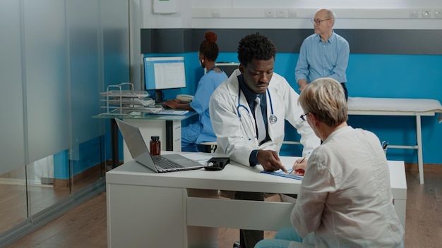 Adulto trabalhando como médico de jaleco branco conversando com paciente velho na mesa no gabinete médico. Mulher mais velha doente usando caneta para assinatura em arquivos de exame sentado com jovem especialista