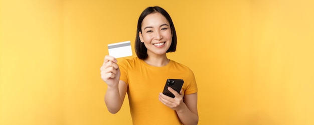 Alegre menina asiática sorrindo mostrando cartão de crédito e smartphone recomendando suporte bancário para celular