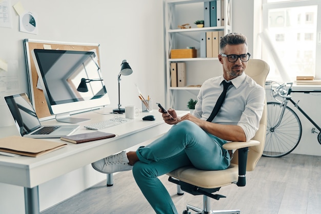 Anstrengender Tag. Gut aussehender junger Mann in Hemd und Krawatte mit Smartphone und lächelnd beim Sitzen im Büro sitting