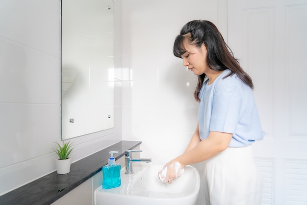 Asiatische Frau, die Hände mit Seife unter dem Wasserhahn mit Wasser im Badezimmer zu Hause wäscht. Stoppen Sie den Covid-19-Ausbruch