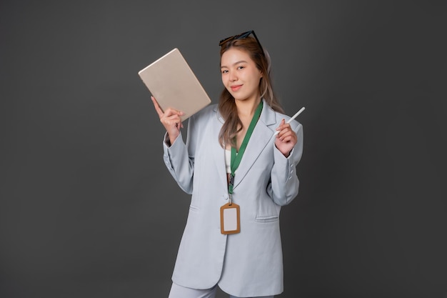 Foto asiatische führungskraft mit langen haaren oder mitarbeiterin eines unternehmens, die einen blauen anzug trägt, eine modebrille trägt, lächelnd ein tablet und einen stift in der hand hält, hängt eine mitarbeiter-id-karte auf, macht ein foto im grauen hintergrund des studios