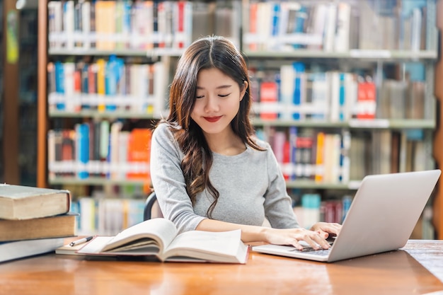 Foto asiatischer junger student in der zufälligen klage, die hausarbeit tut und technologielaptop in der bibliothek der universität verwendet