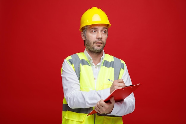 Bauarbeiter in Bauuniform und Schutzhelm mit Klemmbrett, der mit ernstem Gesicht zur Seite schaut und Notizen macht, die über rotem Hintergrund stehen