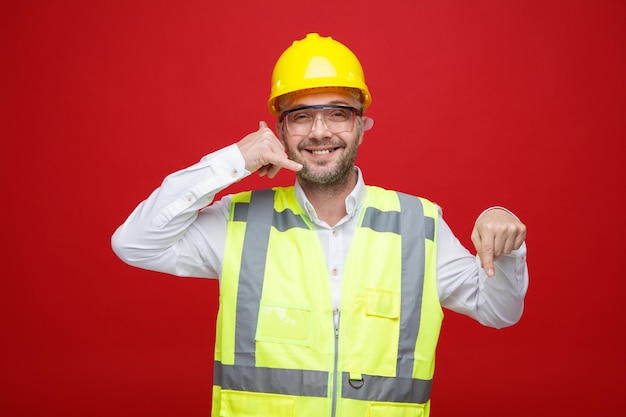 Bauarbeiter in Bauuniform und Schutzhelm mit Schutzbrille, der fröhlich lächelnd in die Kamera blickt und mit dem Zeigefinger nach unten über rotem Hintergrund steht