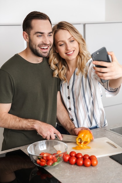 Bild eines glücklichen jungen Liebespaares, das zu Hause in der Küche posiert und sich umarmt, macht ein Selfie mit dem Handy.