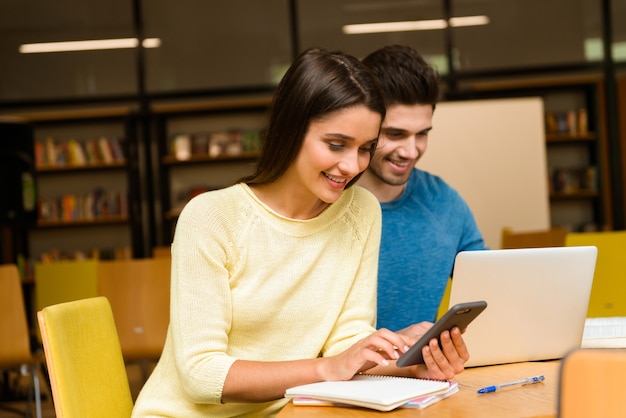 Bild eines jungen Studentenfreundepaares in der Bibliothek, die Hausaufgaben macht, die lesen und Laptop und Handy verwenden.