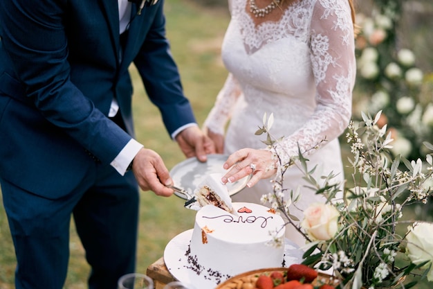 Foto braut und bräutigam legen ein stück hochzeitstorte auf einen teller, der gesichtslos geschnitten ist.