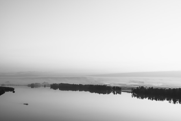 Breiter Fluss fließt entlang diagonales Ufer mit Schattenbild des Waldes und des starken Nebels in Graustufen.