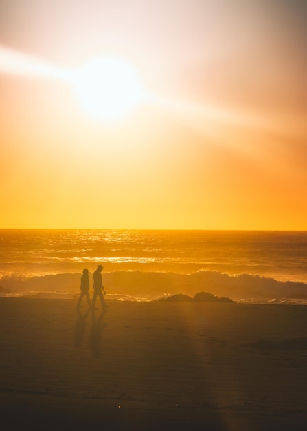 Foto casal de silhuetas a caminhar na praia contra o céu laranja