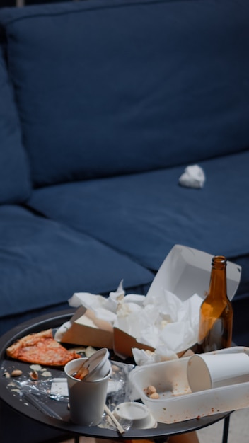 Chaos im leeren Wohnzimmer mit Essensmüll, Flasche Bier und Servietten auf blauem Sofa