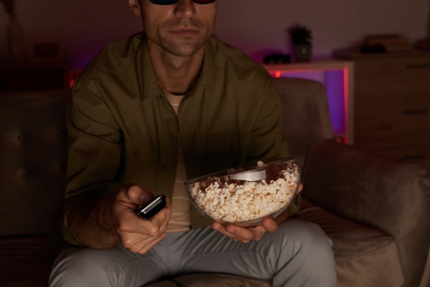 Foto close de jovem segurando a tigela com pipoca e controle remoto e assistindo tv no sofá em casa