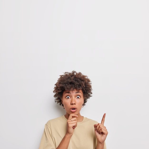 Conceito de promoção. Mulher de cabelos encaracolados surpresa aponta o dedo indicador acima oferece grande venda mostra espaço em branco para o design do seu banner chocou a expressão vestida casualmente posa contra a parede branca