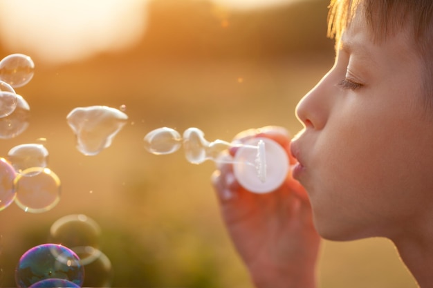 Criança soprando bolhas de sabão ao pôr do sol em um dia de verão. Menino brincando na natureza.