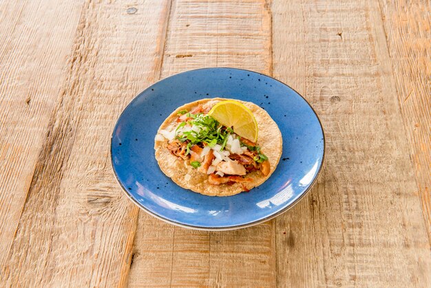 Foto delicioso taco mexicano de carnitas com carne cozida de coentro com cebola crua e rodela de limão com tortilha de trigo e abacate azul na bela mesa de madeira clara