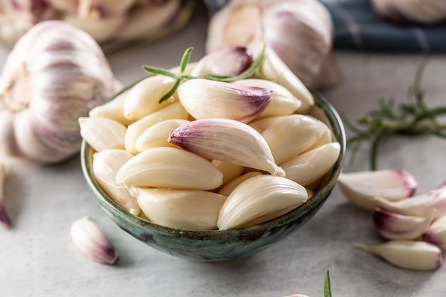 Dentes de alho aromáticos recheados em tigela rústica azul com saboroso alecrim como decoração e mais alho ao fundo.