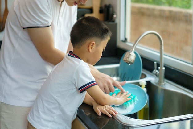 Der asiatische Vater bringt seinem Sohn das Geschirrspülen bei, hilft bei der Hausarbeit in der Küche zu Hause, Väter interagieren den ganzen Tag mit ihren Kindern.