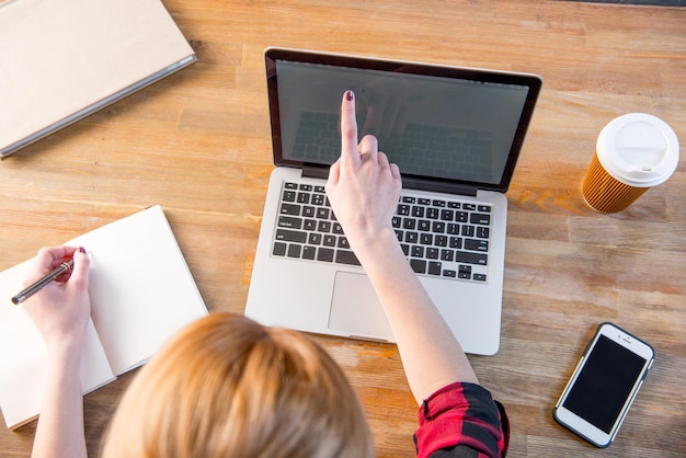 Draufsicht der jungen Frau, die Laptop verwendet und Notizen im Notizbuch macht