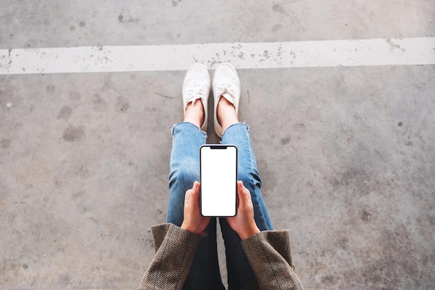 Draufsichtmodell einer Frau, die ein schwarzes Mobiltelefon mit leerem weißem Bildschirm hält, während sie auf dem Boden sitzt