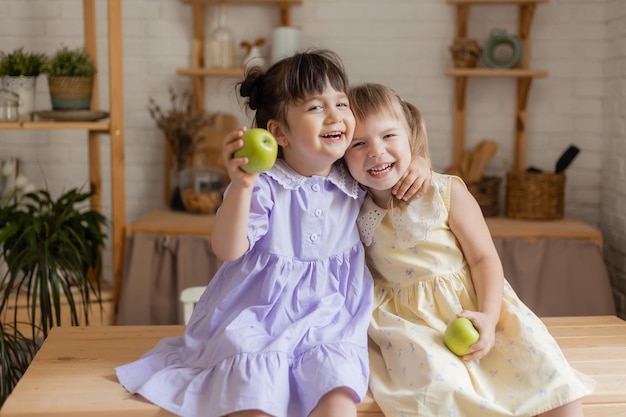 Foto duas meninas engraçadas bonitinhas em vestidos comendo maçãs verdes na cozinha. espaço para texto, banner