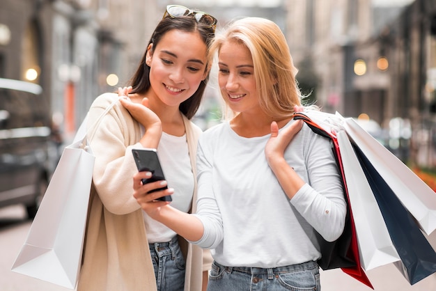 Duas mulheres ao ar livre, olhando para o smartphone, segurando sacolas de compras