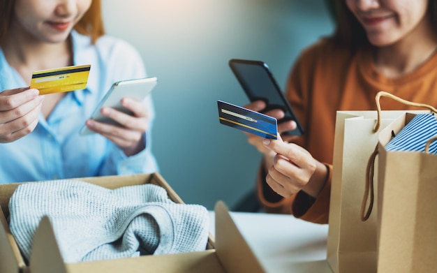 Foto duas mulheres usando telefone celular e cartão de crédito para fazer compras online com sacola de compras e caixa postal com roupas na mesa