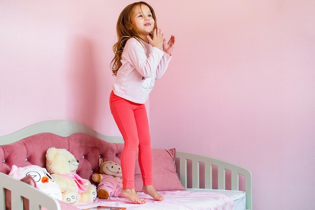 Ein kleines fröhliches Mädchen mit langen blonden Haaren springt in ihrem Zimmer auf ein gemütliches Kinderbett