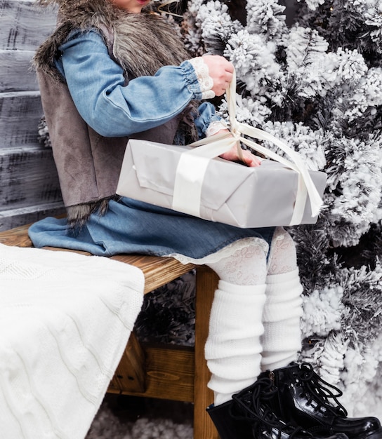 Foto ein kleines mädchen in einem blauen kleid, ärmellosen jacken und weißen strumpfhosen sitzt auf einer bank im neujahrsdekor und öffnet ein neujahrsgeschenk. foto