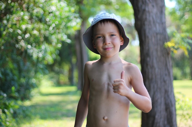 Foto ein lächelnder weißer schuljunge ohne kleidung hält im sommer vor dem hintergrund einer tropischen landschaft den daumen hoch. toller sommerurlaub für die familie