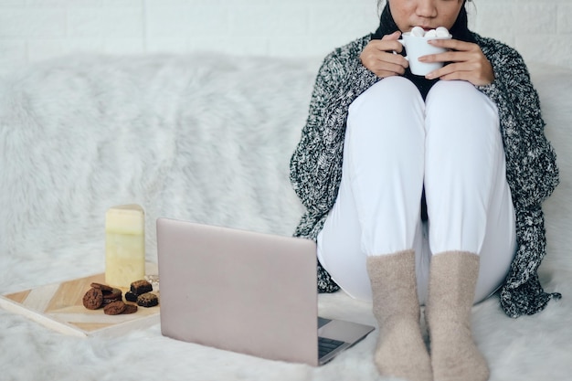 Ein Mädchen mit Winterkleidung sitzt und trinkt heiße Schokolade, während es zu Hause mit dem Laptop arbeitet