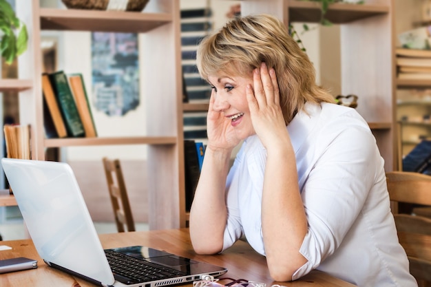 Eine Frau im Büro arbeitet an einem Laptop. Sie schaut auf den Bildschirm und ist glücklich. Eine Frau mittleren Alters, ein Erwachsener in einem Café.