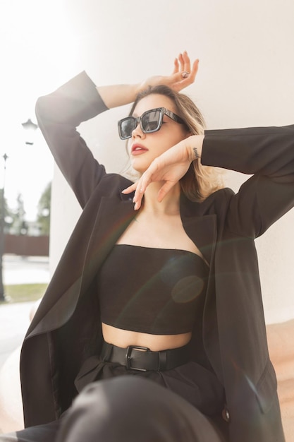 Elegante na moda jovem elegância modelo de mulher bonita com roupa de voga de negócios preto com top blazer e calça senta-se e posa perto de uma coluna branca ao pôr do sol