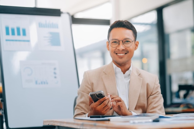 Foto empreendedor asiático excitado usando smartphone e laptop trabalhando online enquanto está sentado no local de trabalho no escritório em casa empresário enviando mensagens de texto por celular e sorrindo