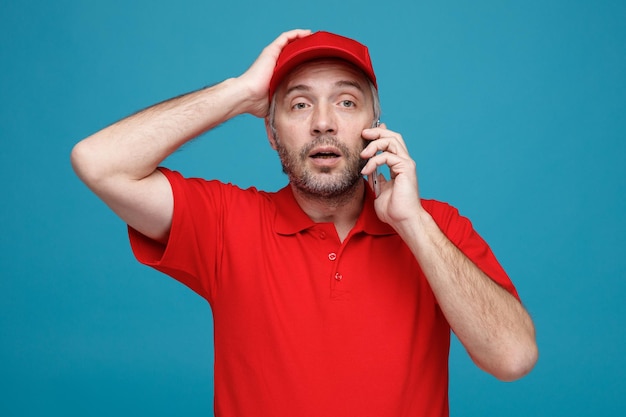 Empregado de entregador em uniforme de camiseta em branco de boné vermelho falando no celular parecendo confuso com a mão na cabeça por erro em pé sobre fundo azul