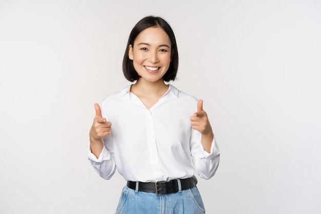 Es ist Ihnen herzlichen Glückwunsch Lächelnde, attraktive asiatische Geschäftsfrau, die mit dem Finger auf die Kamera zeigt, mit zufriedenem Gesicht, das Sie dazu einlädt, vor weißem Hintergrund zu stehen