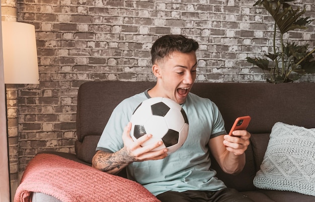 Foto fã de futebol jovem assistindo a um jogo de futebol no celular sentado em um sofá confortável na sala de estar